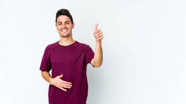 Beyaz arka planda izole edilmiş genç beyaz adam karnına dokunuyor, nazikçe gülümsüyor, yemek yiyor ve tatmin oluyor..