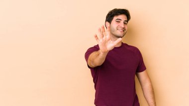 Bej arka planda izole edilmiş genç adam gülümseyerek beş numarayı parmaklarıyla gösteriyor..