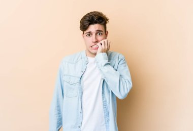 Beyazlı genç bir adam bej arka planda izole edilmiş tırnak yiyor, gergin ve endişeli..