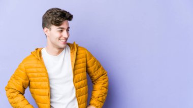 Mor arka planda izole edilmiş genç beyaz adam mutlu bir şekilde gülüyor ve ellerini karnının üstünde tutarak eğleniyor..