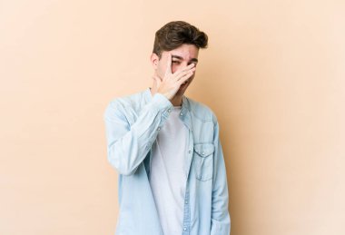 Beyazlı genç bir adam bej arka planda izole edilmiş. Kameraya parmaklarıyla göz kırpıyor, yüzünü kapatmaktan utanıyor..