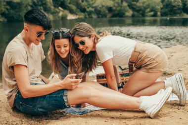 Gülümseyen genç arkadaşlar pikniğin ortasında cep telefonlarını izliyorlar..