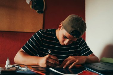 Öğrenci kahverengi şapkalı güneşli odasında notlar alıyor..