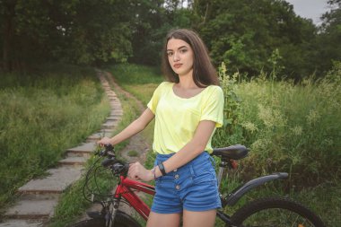 Yol kenarında bisikleti olan genç bir kız. Şortlu ve tişörtlü güzel bir kız dağ bisikletiyle kameraya bakıyor. Yazın çekilen çekimler..