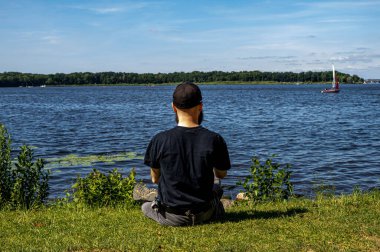 Yalnız sakallı bir adam göldeki yeşil çimlerde oturuyor ve tekneye bakıyor.. 