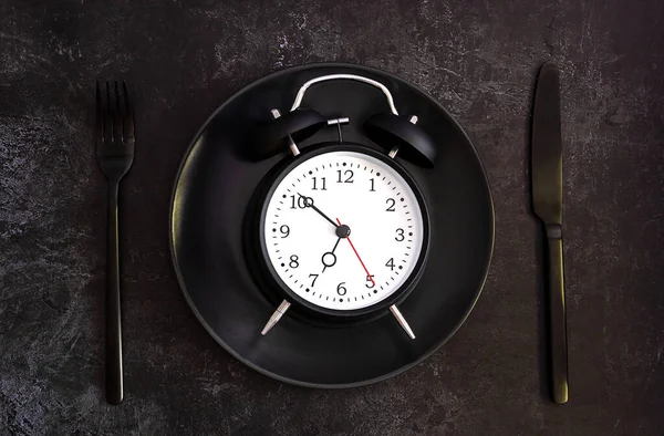 Black alarm clock on a black plate with black knife and fork.