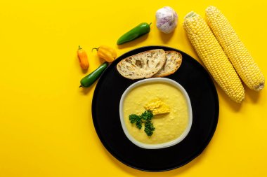 Sarı zemin üzerinde siyah bir tabakta sonbahar baharatlı mısır çorbası. Mısır koçanı ve kızarmış ekmekle servis edilir. Vejetaryen tarifi. 