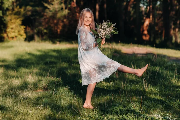 Ein schönes neunjähriges blondes Mädchen mit langen Haaren in einem langen weißen Kleid, das einen Strauß Maiglöckchen in der Hand hält, spaziert im Park in der Natur. Sommer, Sonnenuntergang. — Stockfoto