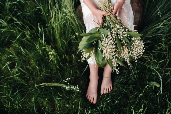 Uma bela loira de nove anos de idade em um vestido branco longo, segurando um buquê de lírios do vale, close-up de uma menina segurando flores do valley.Summer, pôr do sol . — Fotografia de Stock