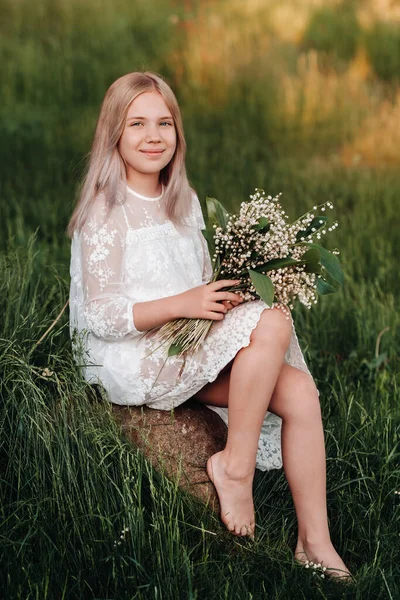 Ein schönes neunjähriges blondes Mädchen mit langen Haaren in einem langen weißen Kleid, das einen Strauß Maiglöckchen in der Hand hält, spaziert im Park in der Natur. Sommer, Sonnenuntergang. — Stockfoto