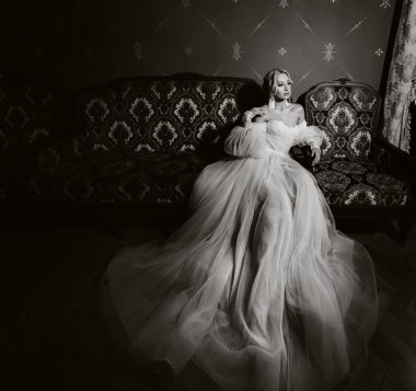 Salonda güzel bir gelinin portresi. Beyaz gelinlikli zarif bir gelin bir sandalyede oturuyor. Siyah beyaz fotoğraf..