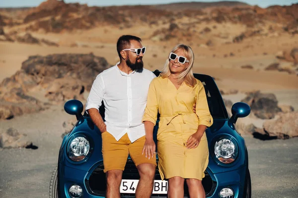 a stylish couple, a man and a woman, stand next to a blue car against the backdrop of the desert on the island of Tenerife. beautiful man and woman in orange dress in the Canary Islands.Spain.The Teide Volcano.