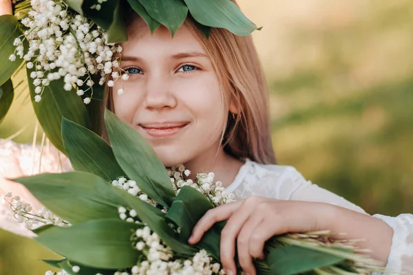 長い白いドレスに長い髪をした美しい9歳のブロンドの女の子は 渓谷の花のユリの花束を持ち 公園で自然の中を歩く — ストック写真