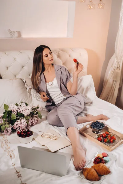 a girl sits in bed in the evening watching a laptop and eating strawberries, a girl in bed eats sweets before going to bed.