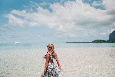 Mayolu bir kız okyanusta duruyor ve Mauritius adasında Le Morne Dağı 'nın arka planında bir deniz uçağı bekliyor. Sudaki bir kadın Mauritius adasına inen bir uçağa bakıyor..