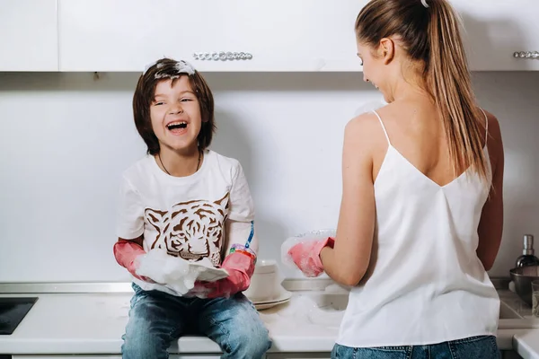 家庭主妇妈妈戴着粉色手套 与她的儿子一起用洗涤剂在洗碗槽里洗手 一个穿着白色衣服的女孩和一个戴着石膏的孩子在打扫房子 并用自制的粉色手套洗盘子 一个戴着石膏的孩子在洗盘子 微笑着 — 图库照片