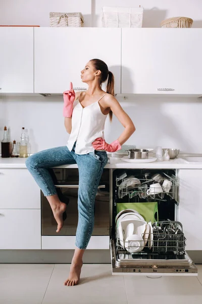 a housewife girl in pink gloves after cleaning the house sits tired in the kitchen.In the white kitchen, the girl has washed the dishes and is resting.Lots of washed dishes.