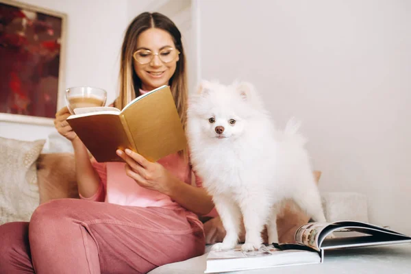 a girl in pajamas at home reads a book with her dog Spitzer, the Dog and its owner are resting on the sofa and reading a book.Household chores.