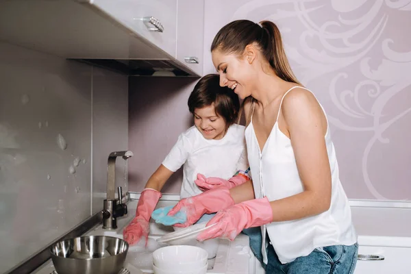 家庭主妇妈妈戴着粉色手套 与她的儿子一起用洗涤剂在洗碗槽里洗手 一个穿着白色衣服的女孩和一个戴着石膏的孩子在打扫房子 并用自制的粉色手套洗盘子 一个戴着石膏的孩子在洗盘子 微笑着 — 图库照片