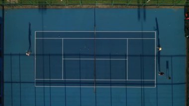 Mavi tenis kortunun en üst görüntüsü. Oyuncular antrenmanda. Akşam vakti sokakta insanlarla tenis kortu.