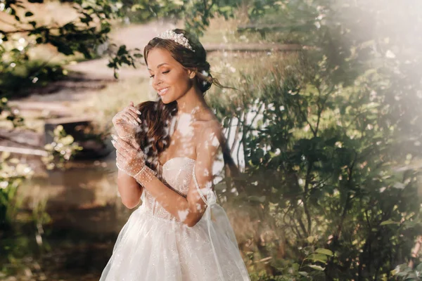 An elegant bride in a white dress, gloves with a bouquet on a waterfall in the Park, enjoying nature.Model in a wedding dress and gloves in the forest.Belarus.