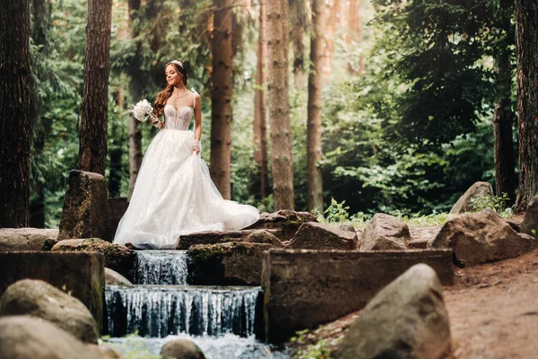An elegant bride in a white dress, gloves with a bouquet on a waterfall in the Park, enjoying nature.Model in a wedding dress and gloves in the forest.Belarus.