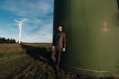 Yeşil golf tişörtlü bir iş adamı, yel değirmeninin yanında, arka planda ve mavi gökyüzünde duruyor. Yel değirmenlerinin yakınında bir iş adamı.