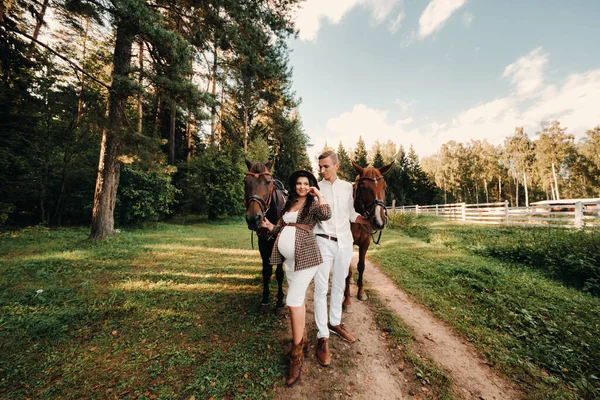 在大自然的森林里 一个戴着帽子的怀孕女孩和她穿着白衣的丈夫站在马旁边 — 图库照片