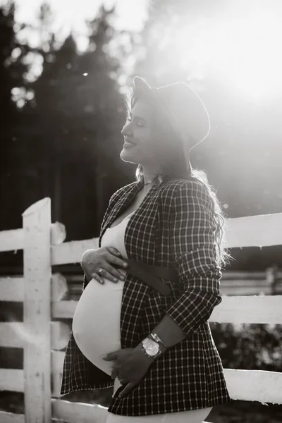 日没時に馬の背の近くの帽子に大きな腹を持つ妊婦 馬と茶色のドレスでスタイリッシュな妊婦 黒と白の写真 — ストック写真