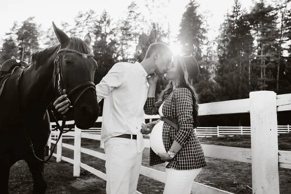 Uma menina grávida em um chapéu e seu marido em roupas brancas ficar ao lado dos cavalos perto do curral cavalo ao pôr do sol.Stylish mulher grávida com um homem com cavalos. Família, preto e branco foto — Fotografia de Stock