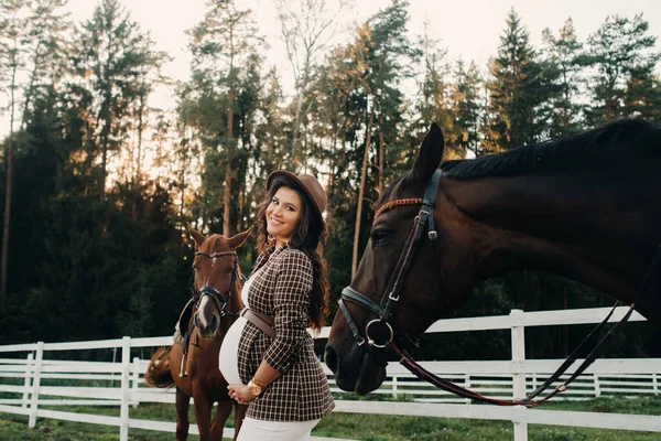 在大自然的围场附近 一个怀孕的女孩 戴着一顶帽子 紧挨着马 穿着棕色连衣裙 穿着马的时髦孕妇 — 图库照片