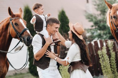 Beyaz elbiseli bir aile ve oğulları doğada iki güzel atın yanında dururlar. Çocuklu şık bir çift atlarla fotoğraf çekilir.