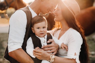 Beyaz elbiseli bir aile ve oğulları doğada iki güzel atın yanında dururlar. Çocuklu şık bir çift atlarla fotoğraf çekilir.