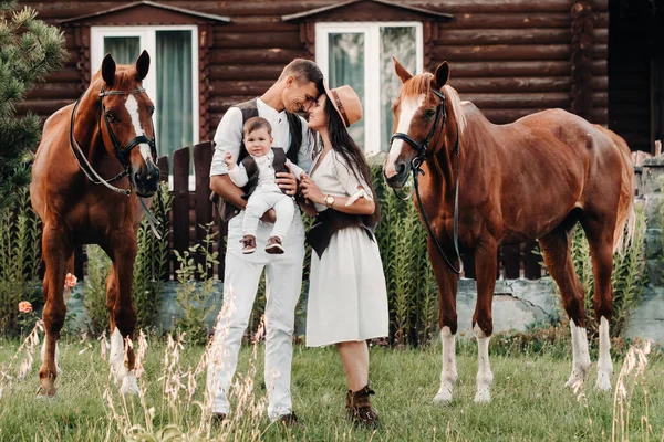 A family in white clothes with their son stand near two beautiful horses in nature. A stylish couple with a child are photographed with horses.