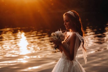 Beyaz elbiseli ve eldivenli zarif bir gelin gün batımında doğanın tadını çıkaran bir buketle birlikte parkta nehrin kenarında duruyor. Bir gelinlik ve eldivenli bir manken..