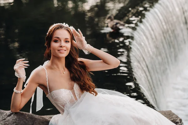 An elegant bride in a white dress, gloves and bare feet is sitting near a waterfall in the Park enjoying nature.A model in a wedding dress and gloves at a nature Park.Belarus.