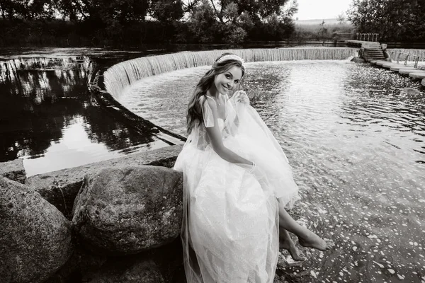 An elegant bride in a white dress, gloves and bare feet is sitting near a waterfall in the Park enjoying nature.A model in a wedding dress and gloves at a nature Park.Belarus.black and white photo