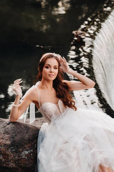 An elegant bride in a white dress, gloves and bare feet is sitting near a waterfall in the Park enjoying nature.A model in a wedding dress and gloves at a nature Park.Belarus.