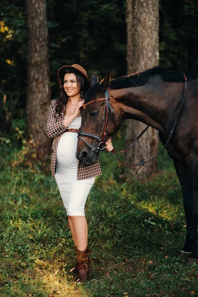 自然の中で馬の隣の帽子に大きな腹を持つ妊娠中の女の子白い服と茶色のジャケットのスタイリッシュな女の子 — ストック写真