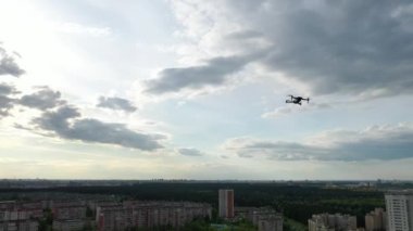 Uçan bir dron bulutlu bir gökyüzüne ve ormanın arka planına doğru havalanıyor. Kuadkopter şehrin ormanlık ve yerleşim bölgesine doğru uçuyor.