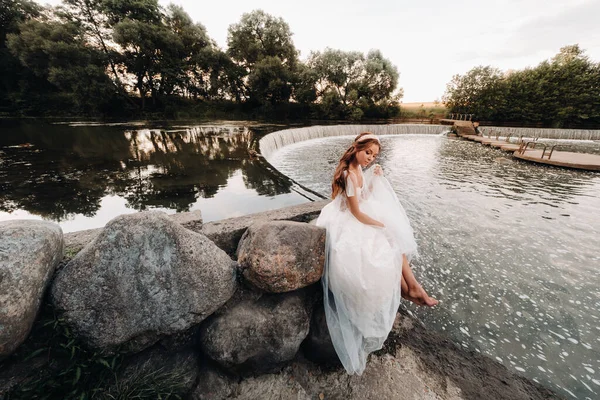 An elegant bride in a white dress, gloves and bare feet is sitting near a waterfall in the Park enjoying nature.A model in a wedding dress and gloves at a nature Park.Belarus.