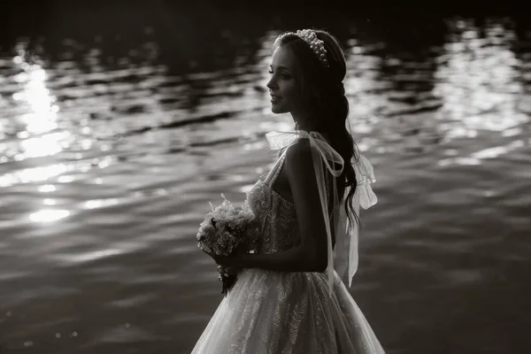 An elegant bride in a white dress and gloves stands by the river in the Park with a bouquet, enjoying nature at sunset.A model in a wedding dress and gloves in a nature Park.Belarus. black and white photo