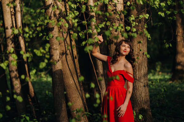 Portrait of a young beautiful laughing girl with long brown hair, in a long red dress in nature, around flowering trees the season is spring.