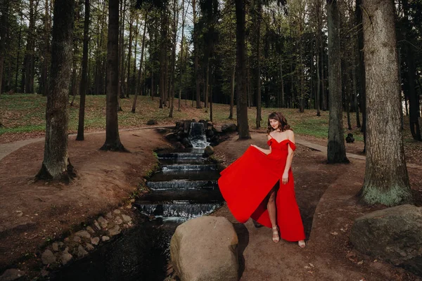 長い赤のドレスで 長い茶色の髪を持つ若い美しい少女の肖像画は 今年の時間 公園の小川を女の子が歩いている — ストック写真