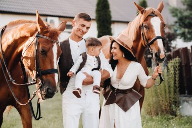 Beyaz elbiseli bir aile ve oğulları doğada iki güzel atın yanında dururlar. Çocuklu şık bir çift atlarla fotoğraf çekilir..