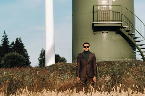 Yeşil golf tişörtlü bir iş adamı, yel değirmeninin yanında, arka planda ve mavi gökyüzünde duruyor. Yel değirmenlerinin yakınında bir iş adamı.