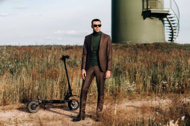 Takım elbiseli ve yeşil golf tişörtlü bir adam elektrikli scooterın yanında mavi gökyüzüne karşı duruyor. Motosikletin yanında iş adamı. Modern gelecek kavramı.