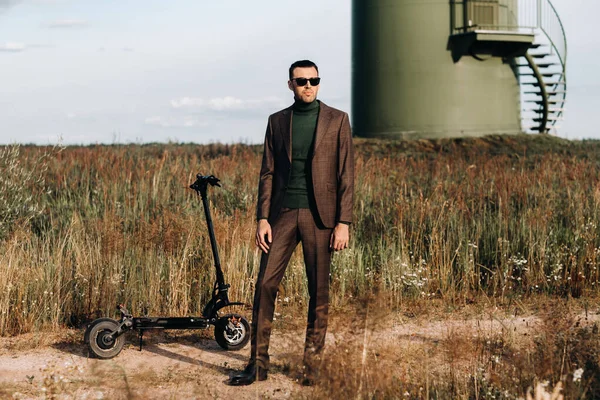 Takım elbiseli ve yeşil golf tişörtlü bir adam elektrikli scooterın yanında mavi gökyüzüne karşı duruyor. Motosikletin yanında iş adamı. Modern gelecek kavramı.