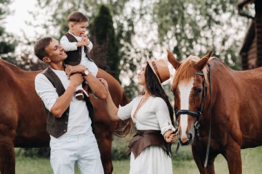 Beyaz elbiseli bir aile ve oğulları doğada iki güzel atın yanında dururlar. Çocuklu şık bir çift atlarla fotoğraf çekilir..