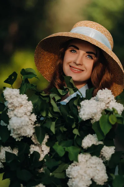 ストローハットのスタイリッシュな女性は サニースプリングパークのライラックの花にポーズをとっています 春の庭にライラックの花束で立っている美しい女の子の穏やかな肖像画 — ストック写真